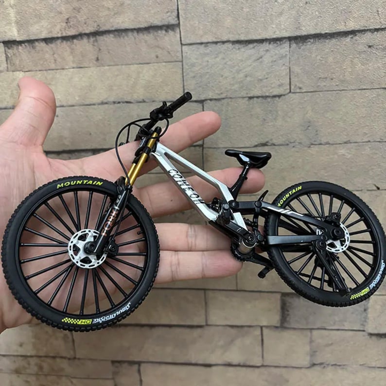 May include: A miniature mountain bike with a silver frame, black tires, and gold suspension. The tires have the word "MOUNTAIN" in yellow. The bike is held in a hand against a brick wall background.