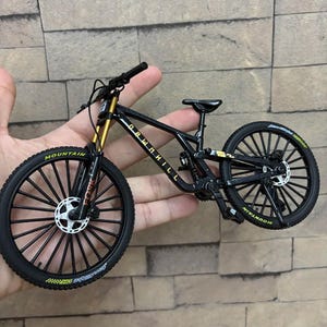Puede incluir: Una bicicleta de monta&ntilde;a en miniatura negra y dorada con la palabra "DOWNHILL" en el cuadro. Los neum&aacute;ticos tienen la palabra "MOUNTAIN" impresa. La bicicleta tiene ruedas negras con radios blancos y horquillas de suspensi&oacute;n doradas.