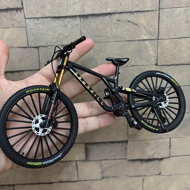 Puede incluir: Una bicicleta de monta&ntilde;a en miniatura negra y dorada con la palabra "DOWNHILL" en el cuadro. Los neum&aacute;ticos tienen la palabra "MOUNTAIN" impresa. La bicicleta tiene ruedas negras con radios blancos y horquillas de suspensi&oacute;n doradas.
