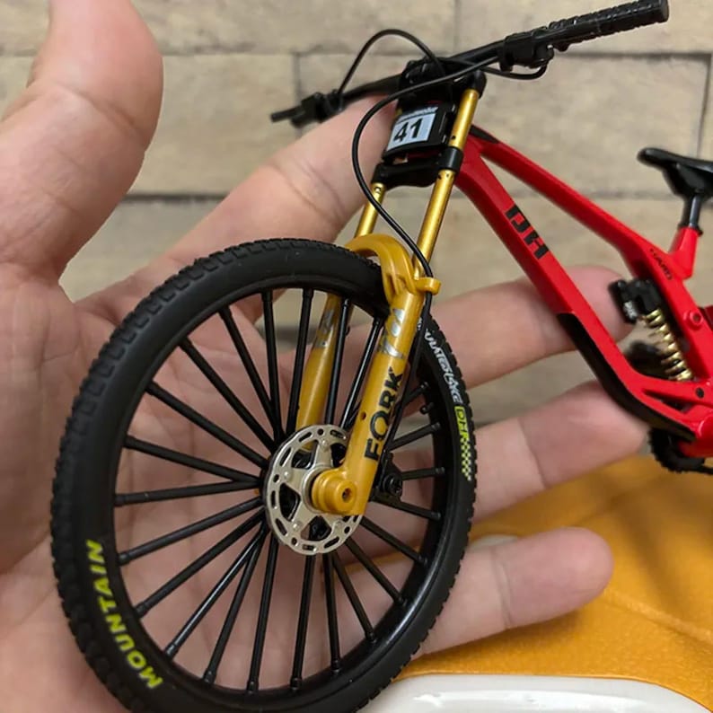 K&ouml;nnte beinhalten: Ein Miniatur-Mountainbike in Rot und Gold mit schwarzen Reifen. Das Vorderrad ist fokussiert und zeigt das Wort "MOUNTAIN" auf dem Reifen. Das Fahrrad hat die Nummer "41" auf dem Rahmen und das Wort "FORK" auf der Vorderradaufh&auml;ngung.