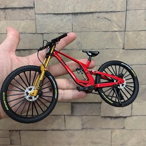 K&ouml;nnte beinhalten: Ein Miniatur-Mountainbike in Rot mit schwarzen Reifen und goldenen Federgabeln. Das Fahrrad hat einen schwarzen Sattel und Lenker. Auf den Reifen steht das Wort "MOUNTAIN". Das Fahrrad wird in der Hand einer Person gehalten.