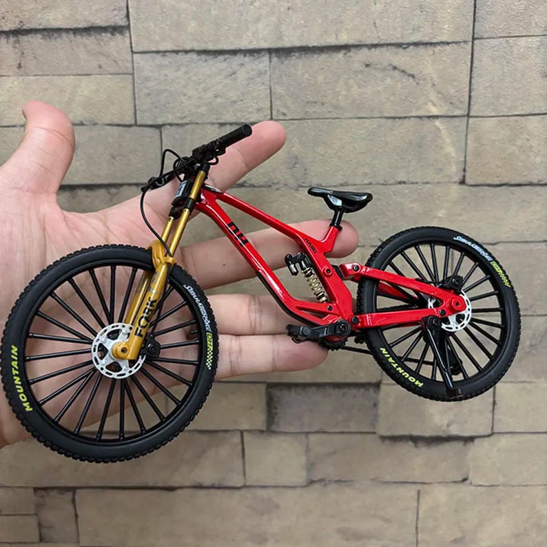 K&ouml;nnte beinhalten: Ein Miniatur-Mountainbike in Rot mit schwarzen Reifen und goldenen Federgabeln. Das Fahrrad hat einen schwarzen Sattel und Lenker. Auf den Reifen steht das Wort "MOUNTAIN". Das Fahrrad wird in der Hand einer Person gehalten.