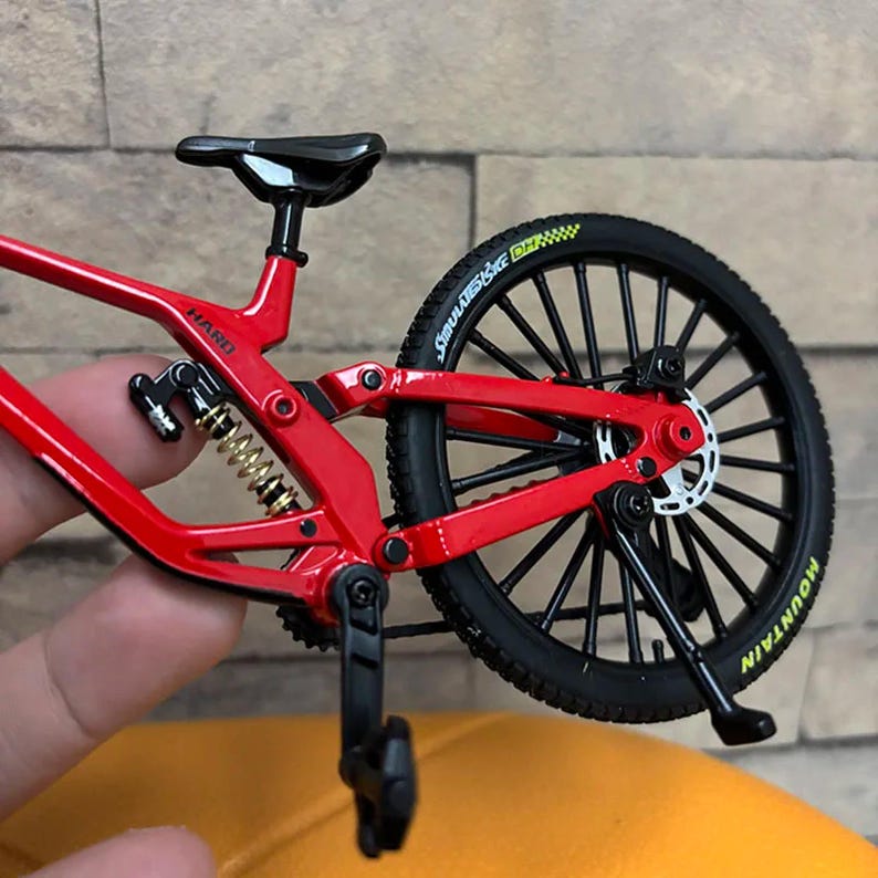 K&ouml;nnte beinhalten: Ein rotes Miniatur-Mountainbike mit schwarzen Reifen und einem schwarzen Sattel. Das Fahrrad hat eine goldene Feder und das Wort "HARD" ist auf dem Rahmen sichtbar. Der Hinterreifen tr&auml;gt das Wort "MOUNTAIN" in Gelb.