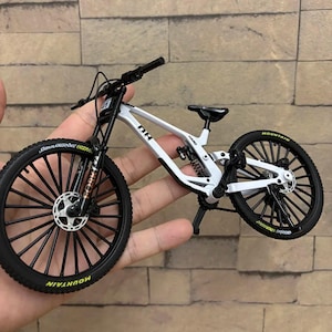 Könnte beinhalten: Ein weiß-schwarzes Miniatur-Mountainbike mit schwarzen Reifen und Rädern. Das Fahrrad hat das Wort "MOUNTAIN" in Gelb auf den Reifen. Das Fahrrad wird in der Hand einer Person gehalten.