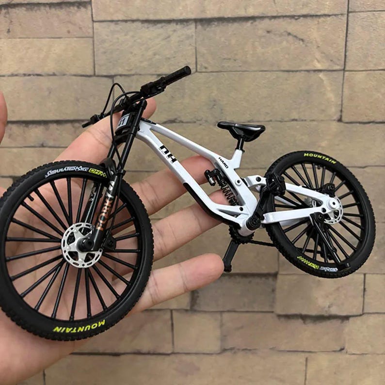 K&ouml;nnte beinhalten: Ein wei&szlig;-schwarzes Miniatur-Mountainbike mit schwarzen Reifen und R&auml;dern. Das Fahrrad hat das Wort "MOUNTAIN" in Gelb auf den Reifen. Das Fahrrad wird in der Hand einer Person gehalten.