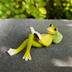 以下が含まれることがあります： 本を読んでいる、リラックスしたライトグリーンのカエルの置物。カエルは白い足、黒い目、緑のクッションをしています。本は白いページと茶色の表紙です。置物は灰色の表面にあります。