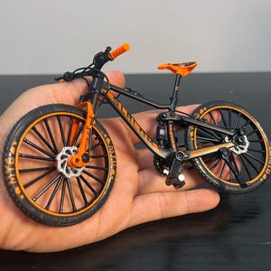 May include: A miniature black and orange mountain bike with orange handlebars, seat, and accents. The bike has black wheels with orange rims and the text "MTB" on the tires. The bike is held in a person's hand.