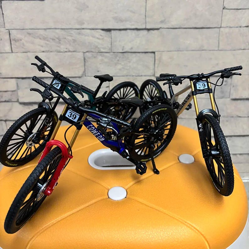 May include: Miniature mountain bikes in various colors, including blue, green, and gold, with black tires and the number 39 on the frame. The bikes are displayed on a yellow surface, showcasing their detailed design and realistic features.