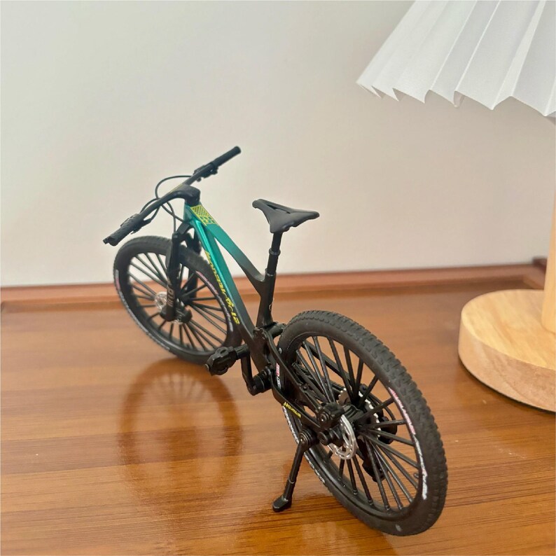 May include: A miniature mountain bike with a black frame and teal and green accents. The bike has black tires, a black seat, and a kickstand. The bike is on a wooden surface.