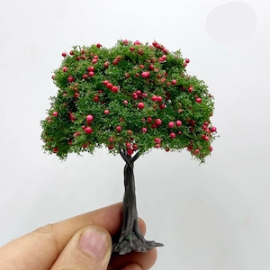 May include: Miniature tree with a dark brown trunk and a full, green canopy dotted with small, red berries. The tree is held by a hand, showcasing its small size and detailed craftsmanship, perfect for model train sets or dioramas.