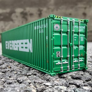 May include: A green Evergreen shipping container model with white text. The container has various markings and logos, including "EMCU 451569 4561". The container is on a textured surface.