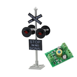 Op de afbeelding: Miniatuur spoorwegovergangssignaal met een zilveren paal, zwart kruisbord en rode lichten. Het bord leest "RAIL CROSSING" en "STOP ON RED SIGNAL". Een groene printplaat staat ook in de afbeelding.