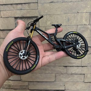 Könnte beinhalten: Ein schwarzes und goldenes Miniatur-Mountainbike mit dem Wort "SIMULATION" auf dem Rahmen. Das Fahrrad hat schwarze Reifen mit dem Wort "MOUNTAIN" in gelben Buchstaben. Das Fahrrad wird in einer Hand gehalten.