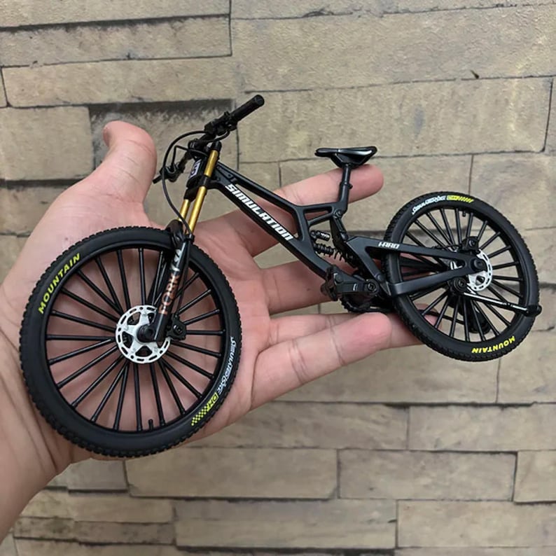May include: A black and gold miniature mountain bike with the word "SIMULATION" printed on the frame. The bike has black tires with the word "MOUNTAIN" in yellow lettering. The bike is held in a hand.