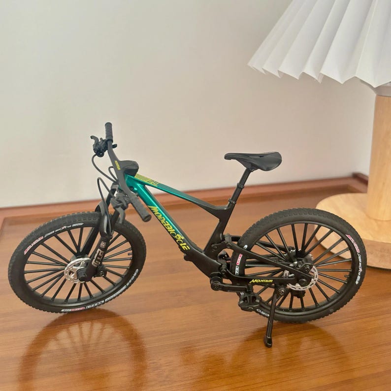 May include: A miniature mountain bike with a black frame and wheels, and a teal and green frame. The bike has a black seat, handlebars, and tires. The text "MODERN CYCLE" is visible on the frame.