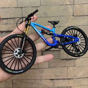 May include: A miniature blue and black mountain bike with gold suspension forks. The bike has black tires and rims, and the words "Down Hill" are visible on the frame. The bike is held in a person's hand.