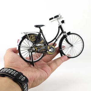 May include: A miniature black bicycle with silver accents and the text "Vintage Shanghai" in gold. The bicycle has a classic design with a rear rack and fenders. The bicycle is held in a person's hand.