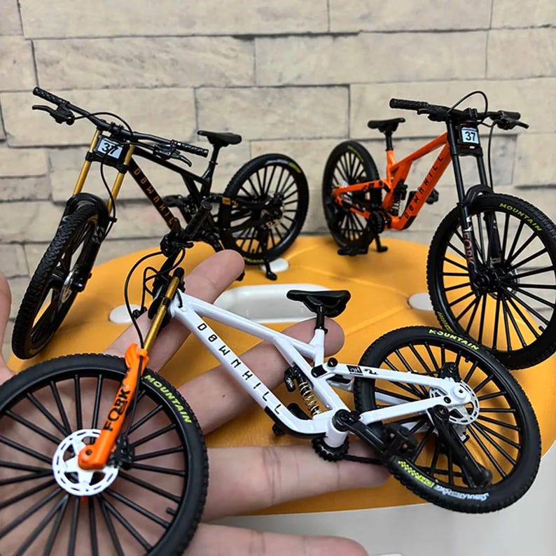 Puede incluir: Mini bicicletas de monta&ntilde;a en negro, blanco y naranja con neum&aacute;ticos negros y la palabra "Downhill" en el cuadro. Las bicicletas tienen el n&uacute;mero 37 en la parte delantera. Las bicicletas est&aacute;n sobre una superficie amarilla.