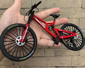 May include: A red and black miniature mountain bike with the word "SIMULATION" printed on the frame. The bike has black tires with the word "MOUNTAIN" printed on the sidewalls. The bike is held in a hand.