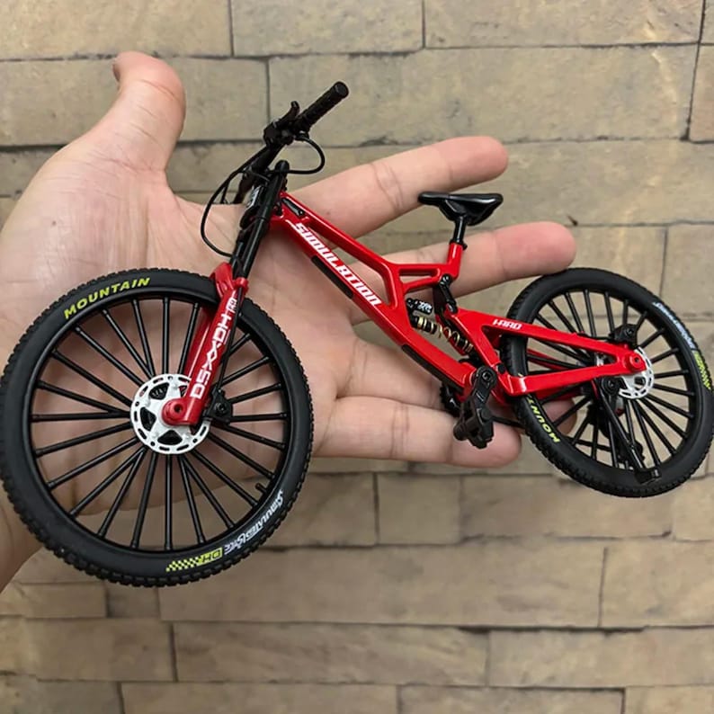 May include: A red and black miniature mountain bike with the word "SIMULATION" printed on the frame. The bike has black tires with the word "MOUNTAIN" printed on the sidewalls. The bike is held in a hand.
