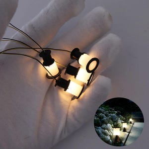 Może przedstawiać: Miniaturowe czarno-białe lampy uliczne z ciepłymi białymi światłami. Lampy są trzymane w rękawicy, z przewodami wychodzącymi z każdej. Mały obrazek wstawiony pokazuje lampy w otoczeniu ogrodu.