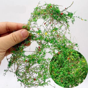 Può includere: Un modello in miniatura dettagliato di un cespuglio verde con minuscoli fiori rossi e gialli. Il modello è tenuto in mano, mostrando la sua delicata costruzione. Ideale per gli appassionati di modellismo ferroviario o per i creatori di diorami.