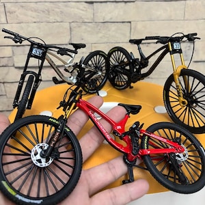 Könnte beinhalten: Miniatur-Mountainbikes in Rot, Schwarz und Silber mit schwarzen Reifen und goldenen Federgabeln. Jedes Fahrrad hat die Nummer 33 am Lenker. Die Fahrräder werden auf einer gelben Oberfläche präsentiert.