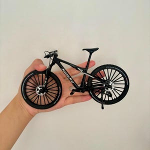 Könnte beinhalten: Ein schwarzes Miniatur-Mountainbike mit dem Text "MODERNCYCLE" auf dem Rahmen. Das Fahrrad hat schwarze Reifen, einen schwarzen Sattel und einen schwarzen Lenker. Das Fahrrad wird in der Hand einer Person gehalten.