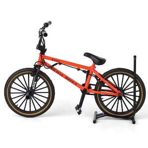 Op de afbeelding: Een oranje BMX-fiets met zwarte banden en velgen. De fiets heeft de woorden "RACE" en "BICYCLE MOTO-X" op het frame gedrukt. Het wordt op een zwarte standaard gepresenteerd.