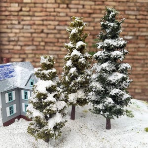 以下が含まれることがあります： 雪に覆われたミニチュアの常緑樹のモデル。背景には小さな灰色の家があります。木はさまざまな緑と茶色の色合いで、白い雪が積もっています。家には屋根にソーラーパネルがあり、白いシャッターが付いています。