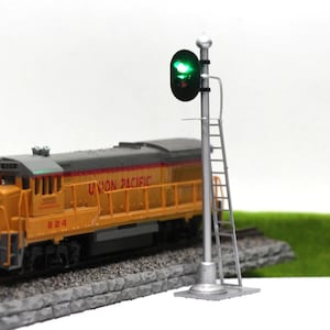 Puede incluir: Una escena de tren en miniatura con una locomotora Union Pacific amarilla y gris y una señal ferroviaria plateada con una luz verde encendida. La señal tiene una pequeña escalera. La locomotora está en una vía.