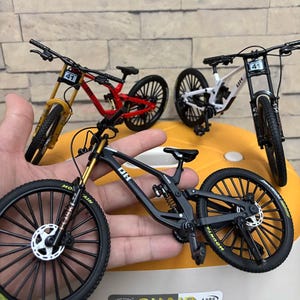 K&ouml;nnte beinhalten: Miniatur-Mountainbikes in Schwarz, Rot, Wei&szlig; und Gelb. Jedes Fahrrad hat detaillierte Merkmale wie Federung, Scheibenbremsen und Reifen. Die Fahrr&auml;der werden auf einer gelben Oberfl&auml;che pr&auml;sentiert.