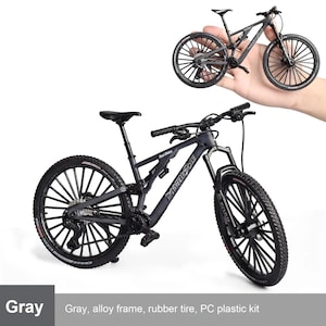 May include: Gray miniature mountain bike with black wheels and tires. The bike has a detailed design, including a seat, handlebars, and suspension. The text "Moda Bicycle" is visible on the frame. The bike is shown in two views.