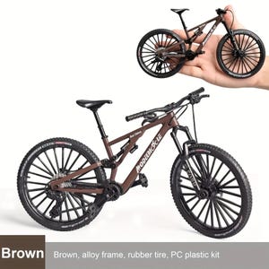 May include: A brown miniature mountain bike with black wheels and tires. The bike has a detailed frame and components, including a seat, handlebars, and suspension. The text "MODEBIOCLE" is visible on the frame. The bike is made of alloy, rubber, and plastic.