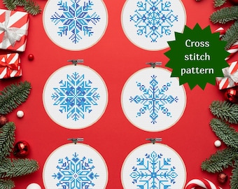 May include: Set of four blue and white snowflake cross stitch patterns in embroidery hoops. The hoops are arranged in two rows of two on a red background. The text "Cross stitch pattern" is in a white speech bubble.
