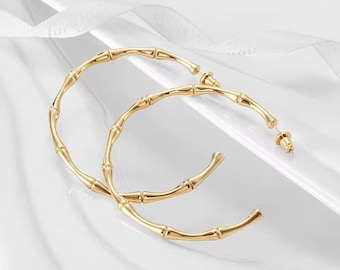 May include: A pair of gold-colored hoop earrings. The earrings have a bamboo-like design, with segmented sections and a curved shape. The earrings are displayed on a white surface with a ribbon-like accent.