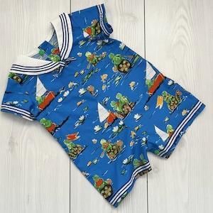 Peut inclure: Barboteuse bleue avec col marin et imprimé fantaisiste de tortues naviguant et ramant sur un fond d'eau bleu. Le col et les manches sont rayés blanc et bleu marine.