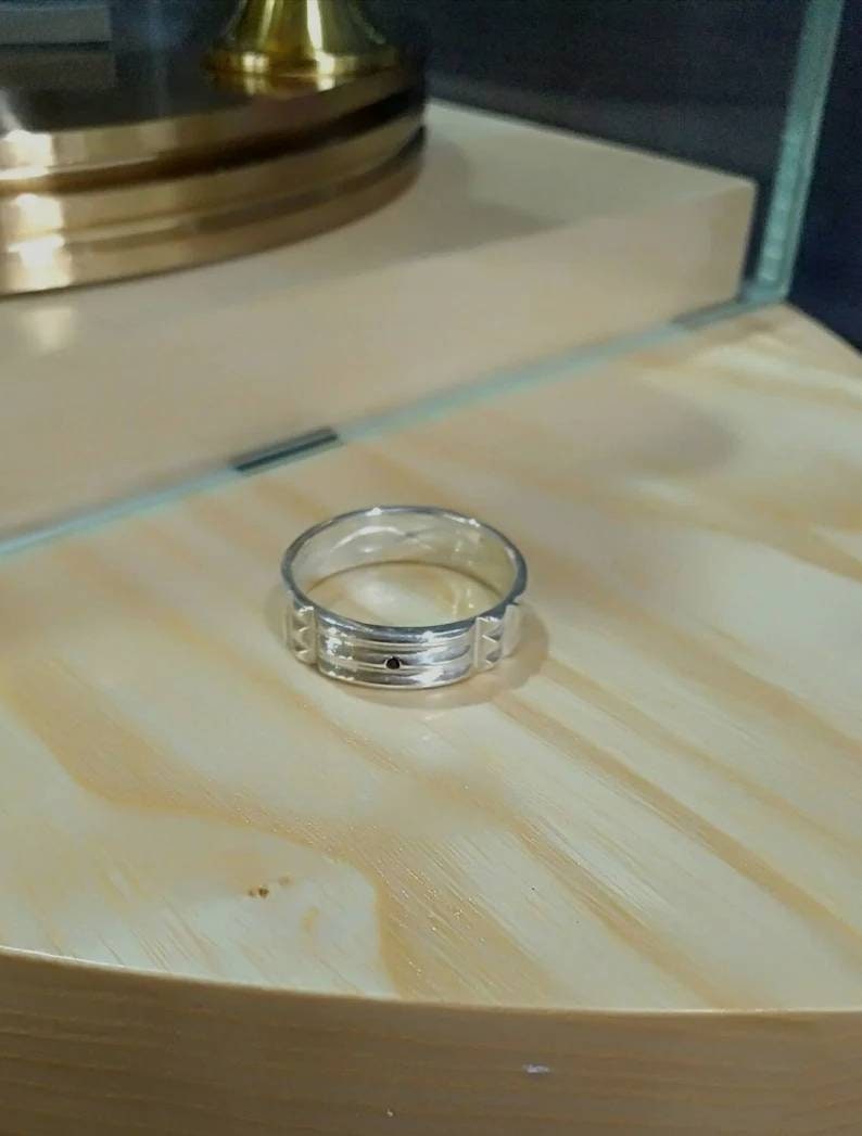 Pu&ograve; includere: Un anello a fascia d'argento con un design testurizzato e una piccola pietra nera al centro.