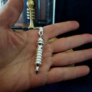Op de afbeelding: Een zilveren metalen slinger met een puntige punt en een spiraalvormig ontwerp. De slinger hangt aan een zwart koord.