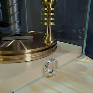 Pu&ograve; includere: Un anello d'argento con un designo testurizzato si trova su una superficie di legno. L'anello &egrave; a fuoco, mentre una scultura in metallo dorato &egrave; sfocata sullo sfondo.