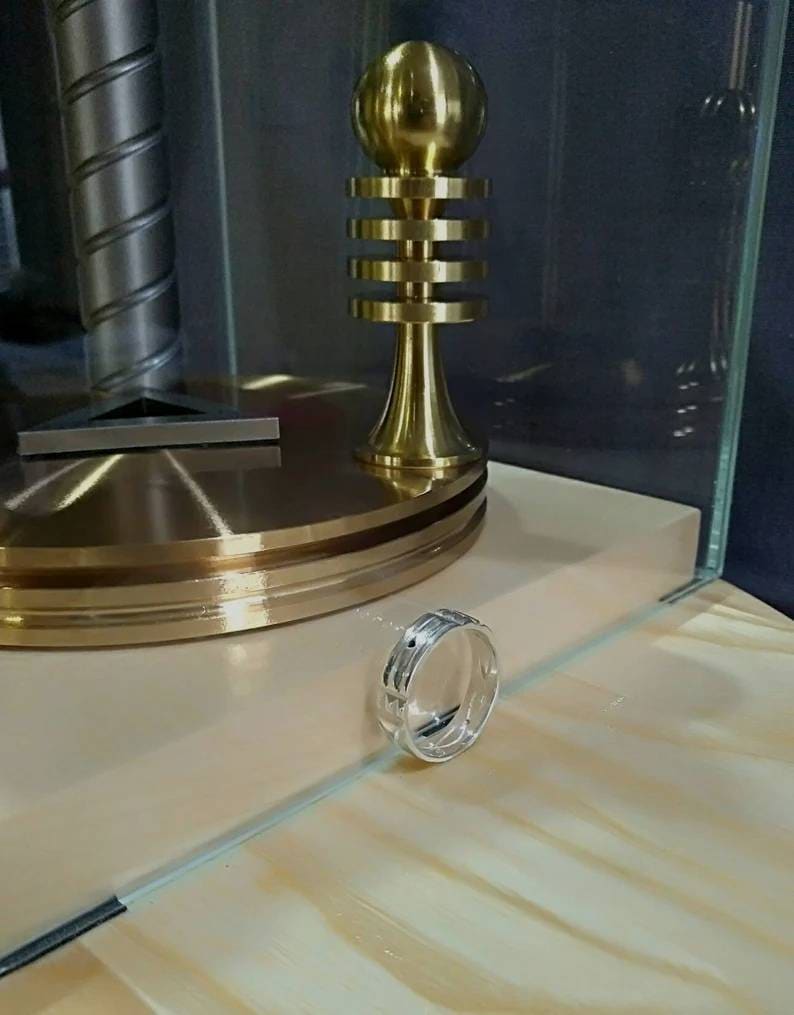 Pu&ograve; includere: Un anello d'argento con un designo testurizzato si trova su una superficie di legno. L'anello &egrave; a fuoco, mentre una scultura in metallo dorato &egrave; sfocata sullo sfondo.