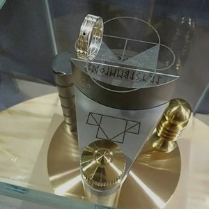 Könnte beinhalten: Ein silberner Ring mit einem schwarzen Stein, der in eine silberne Band gefasst ist. Der Ring befindet sich auf einem Präsentationsständer mit einem goldenen Knopf und einer silbernen Basis. Der Ständer hat einen Kreis mit einem Fadenkreuz und einem Dreieck, das in ein Quadrat eingeschrieben ist.