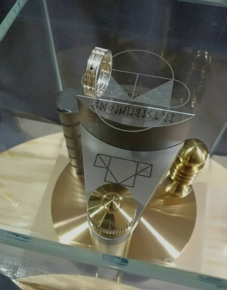 Pu&ograve; includere: Un anello d'argento con una pietra nera incastonata in una fascia d'argento. L'anello &egrave; su un espositore con un pomello dorato e una base d'argento. L'espositore ha un cerchio con un reticolo e un triangolo inscritto in un quadrato.