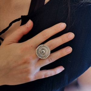 Op de afbeelding: Een zilveren ring met een cirkelvormig ontwerp met een reli&euml;f, getextureerd patroon. De ring wordt aan een vinger gedragen.