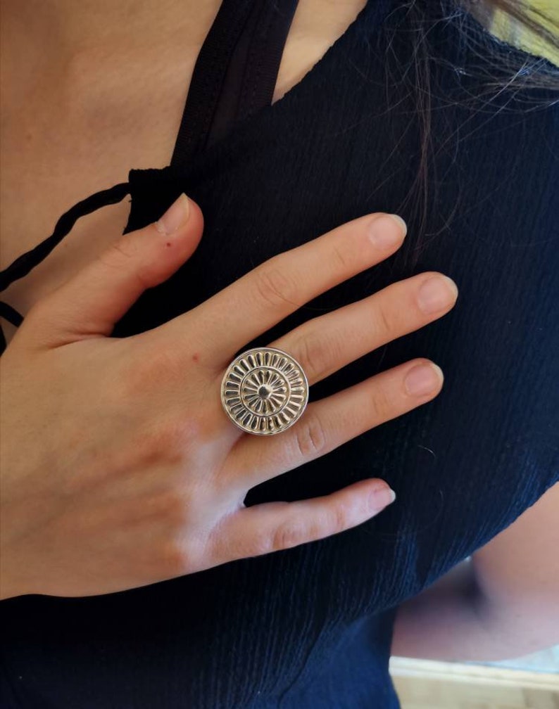 Op de afbeelding: Een zilveren ring met een cirkelvormig ontwerp met een reli&euml;f, getextureerd patroon. De ring wordt aan een vinger gedragen.