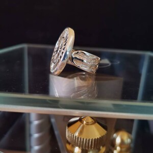 Op de afbeelding: Een zilveren ring met een rond, gedetailleerd ontwerp. De ring ligt op een glazen oppervlak.