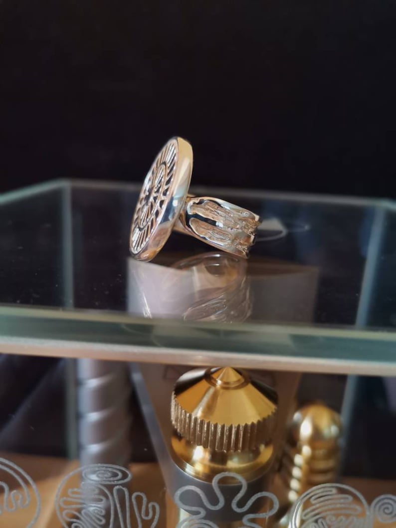 Op de afbeelding: Een zilveren ring met een rond, gedetailleerd ontwerp. De ring ligt op een glazen oppervlak.