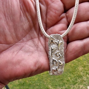 May include: A silver pendant necklace with a textured rectangular charm featuring Hebrew letters. The necklace is on a white cord.