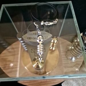 Op de afbeelding: Een zilveren en gouden hangerketting met een zwart koord. De hanger heeft een cilindrisch ontwerp met een cirkelvormige basis en een reeks ribbels. De hanger wordt tentoongesteld in een glazen vitrine met andere metalen objecten.
