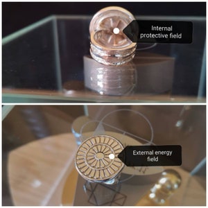 Op de afbeelding: Twee zilveren ringen met een decoratief ontwerp. De bovenste ring heeft een cirkelvormig ontwerp met een verhoogd midden en de onderste ring heeft een band met twee verhoogde lijnen. De ringen liggen op een glazen oppervlak. De bovenste ring heeft tekst die zegt "Intern beschermend veld" en de onderste ring heeft tekst die zegt "Extern energieveld".
